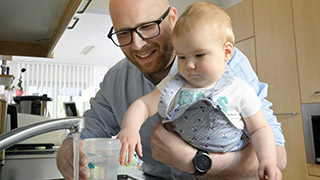 Mann mit Baby auf dem Arm füllt eine Trinkflasche am Wasserhahn
