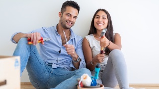 Ein junges Paar sitzt lächelnd mit Werkzeugen am Fußboden einer leeren Wohnung.